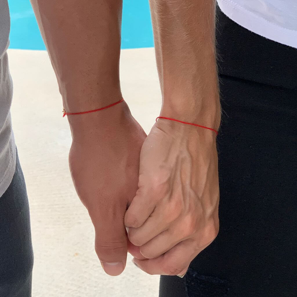 Close-up of two individuals holding hands, showcasing a red string bracelet on one's wrist - Luck Strings