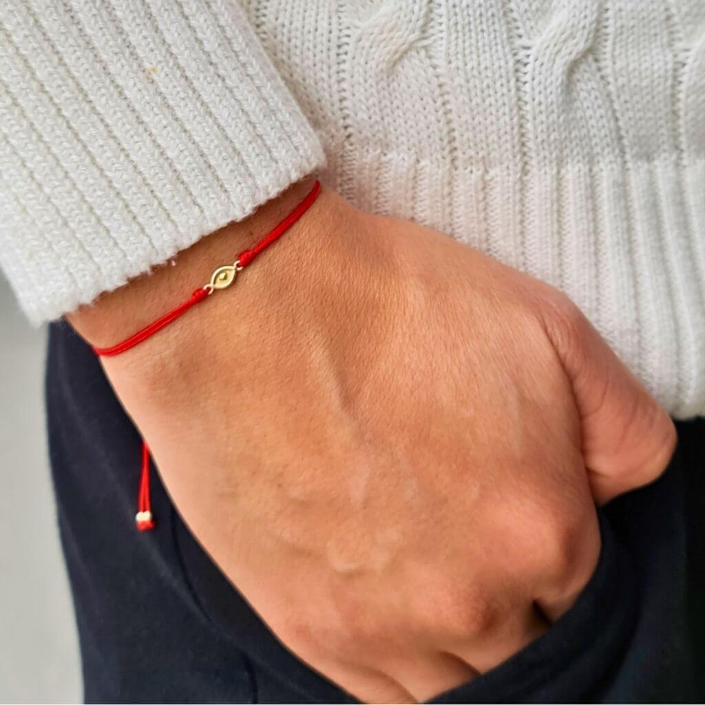 A person's wrist adorned with a simple and elegant red string bracelet, featuring a small, central gold evil eye charm, against the backdrop of a white sweater and black pants - Luck Strings