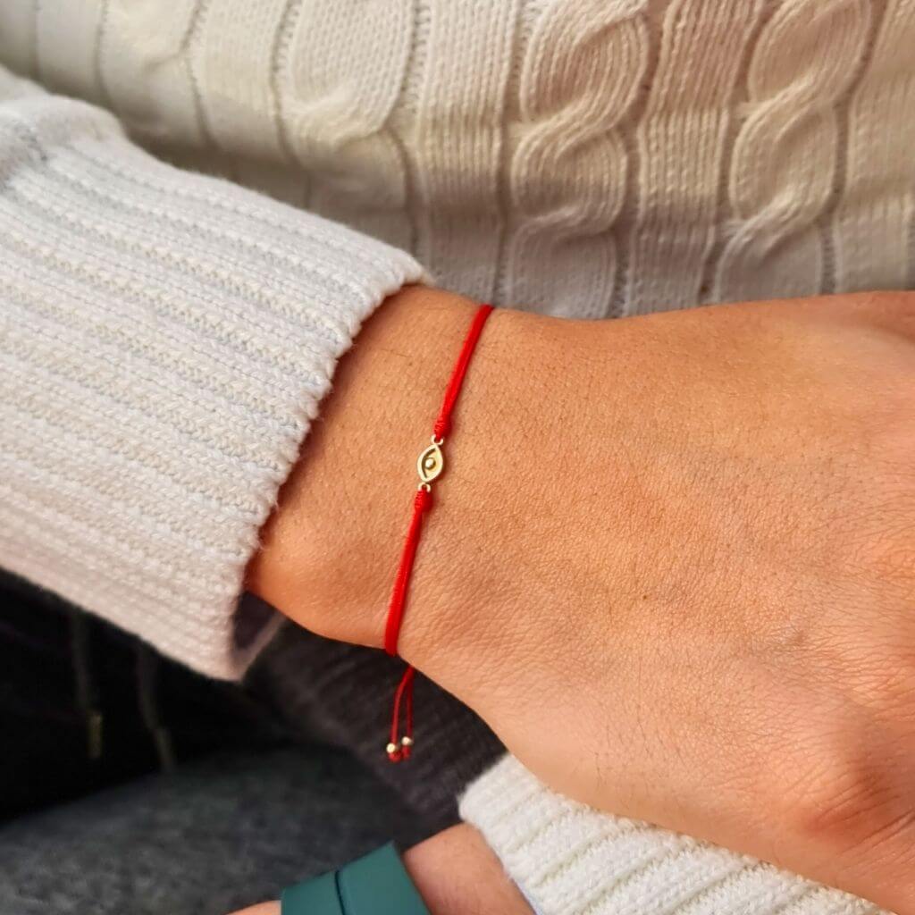 A person&#39;s wrist adorned with a simple and elegant red string bracelet, featuring a small, central gold evil eye charm, against the backdrop of a white sweater and black pants - Luck Strings
